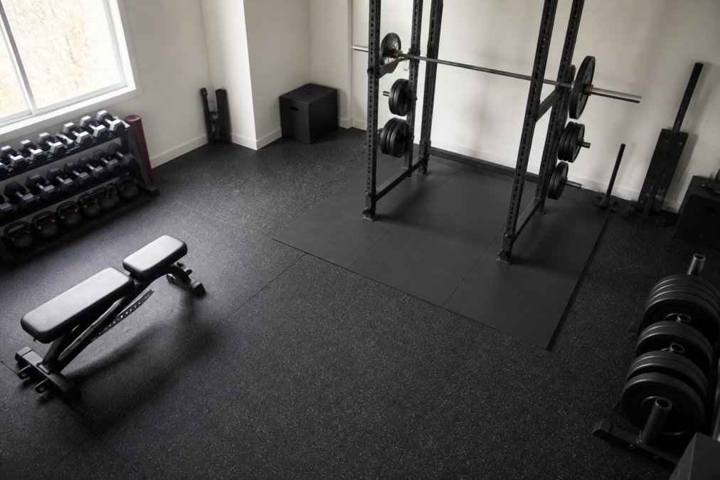 Overhead view of a compact home gym showing power rack positioned with 3-foot clearance on sides, weight bench in separate area, and clear floor path for dynamic movements in a residential room