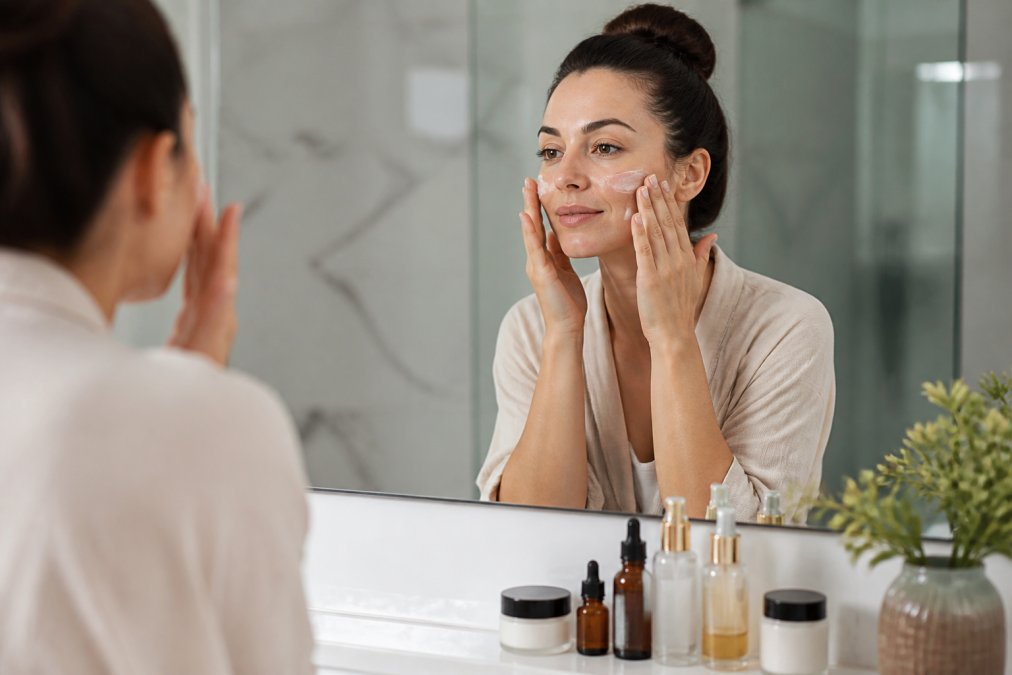 Woman applying hydrating face cream in a bright bathroom mirror reflection