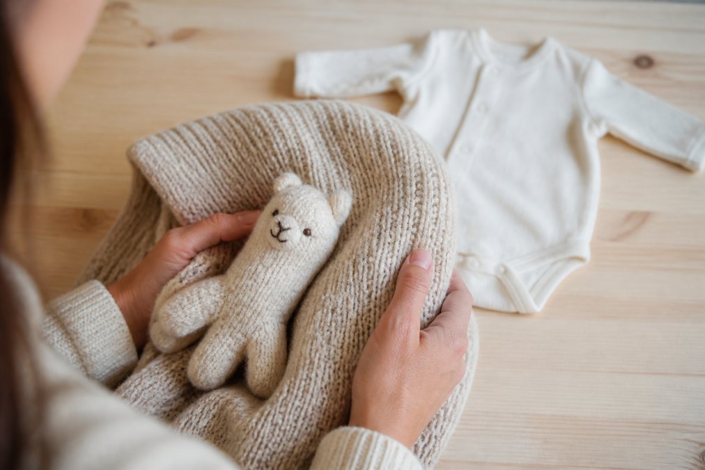 A parent's hands gently holding a soft baby alpaca knit blanket and a small knit toy, with a baby onesie in the background on a light wooden surface