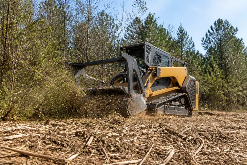 A tracked forestry mulcher machine grinding through dense brush and small trees on a sunny property, leaving a clean layer of wood chips behind