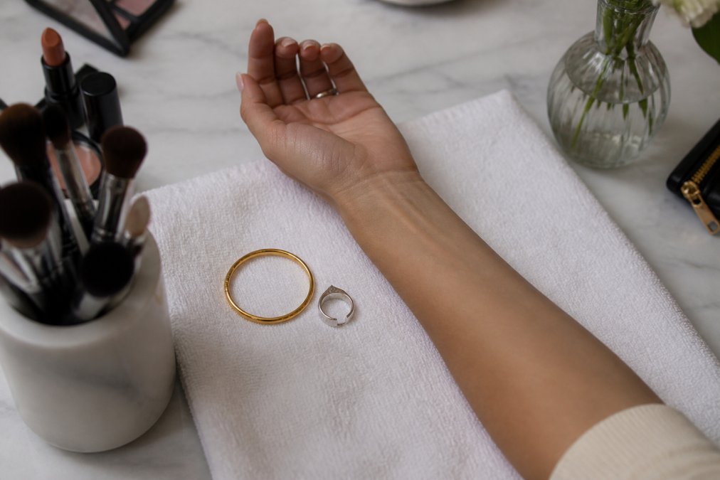 Overhead shot of a woman's forearm resting on a white towel, with a gold bracelet and a silver ring placed beside her wrist to compare which metal complements her skin