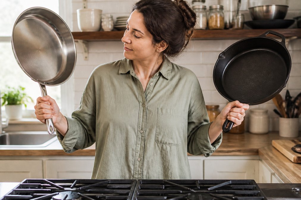 A home cook in a bright kitchen holding a heavy stainless steel frying pan next to a cast iron skillet on a gas stove, showing the weight difference in their hands