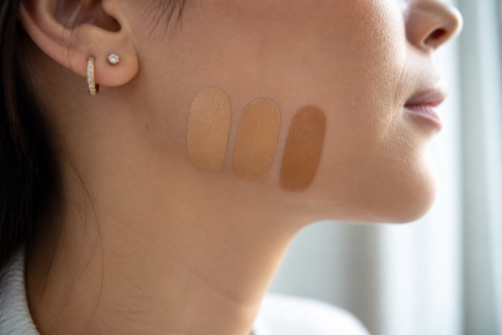 Close-up of a person's jawline with three different foundation swatches visible, comparing shades in natural window light to find the perfect match