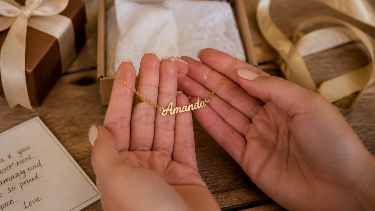 Close-up of hands holding a personalized name necklace with delicate script lettering, positioned on a wooden table next to gift wrapping and a handwritten note