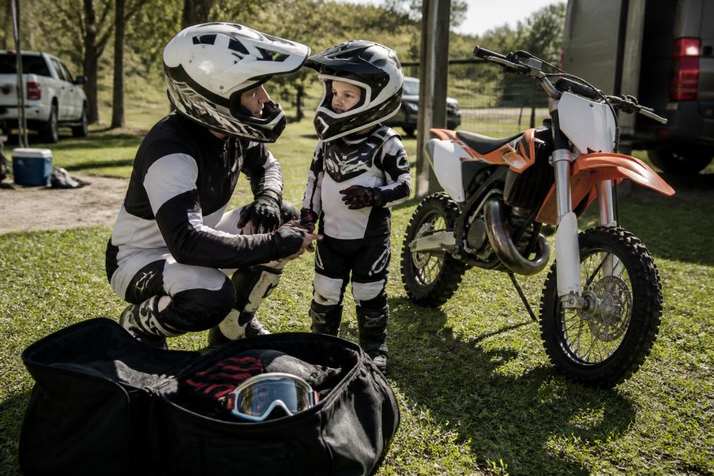 A father and his young child standing beside a dirt bike in a grassy pit area, both wearing motocross jerseys, pants, and helmets, with a gear bag open on the ground showing gloves and goggles