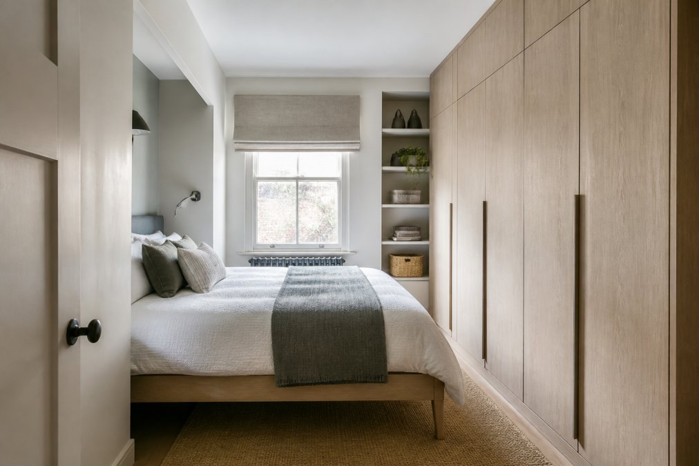 Compact bedroom with custom-fitted wardrobes featuring light wood finish, visible storage solutions in alcoves, bed with raised legs creating visual lightness, and minimalist design with neutral colours creating an organised, clutter-free environment