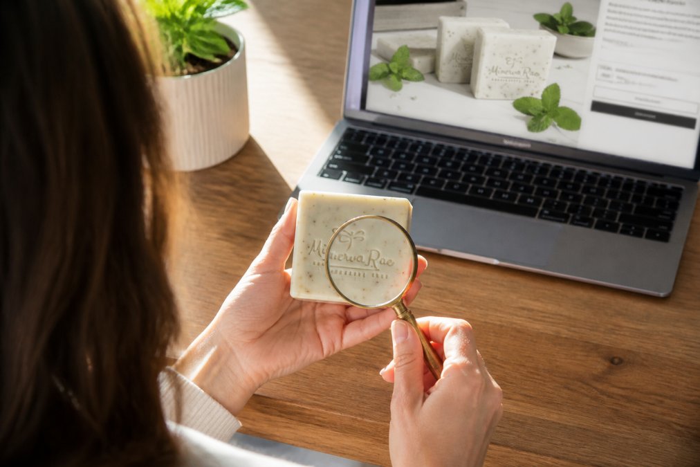 Person sitting at a desk with a laptop open to a soap product page, holding a handcrafted soap bar in one hand while reading the ingredient label with a magnifying glass, with natural sunlight streaming across the wooden desk surface