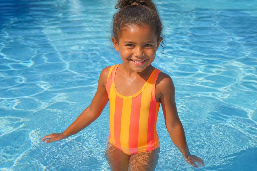 Child wearing a bright neon golden-yellow and coral-striped swimsuit standing in shallow pool water, showing how the vibrant warm neon colors contrast sharply against the blue water while complementing sun-kissed skin