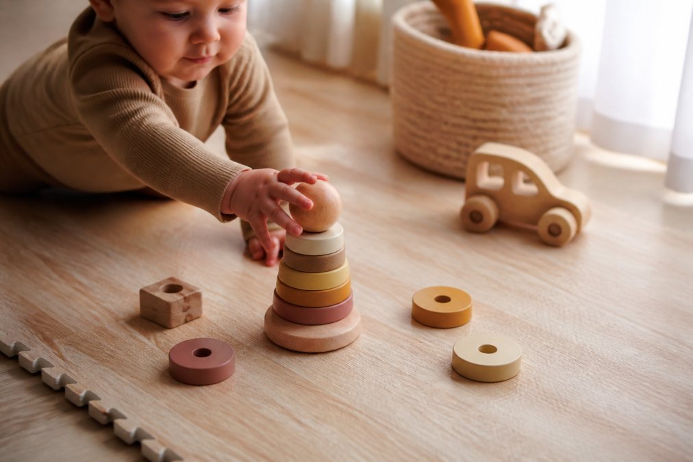 Close-up of a baby's hands reaching for a set of natural wood and silicone stacking toys on a light wooden play mat, soft natural light filtering from a nearby window