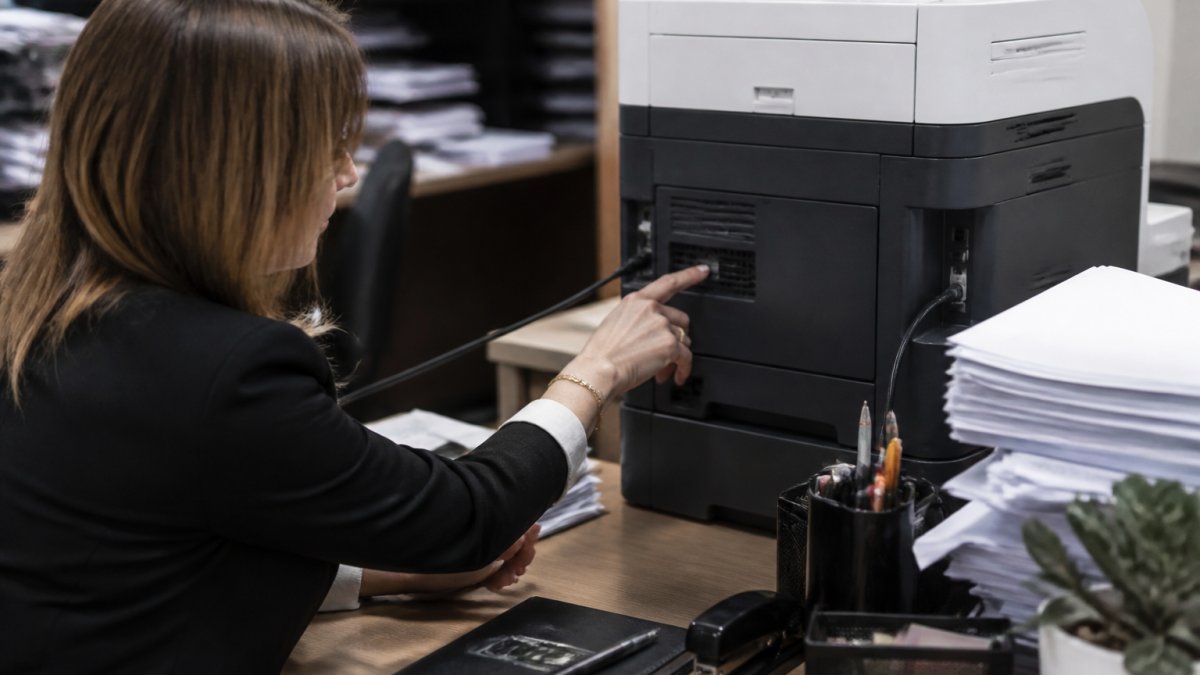 Office manager checking printer model number on the back of a laser printer in a busy office environment with stacks of paper and desk supplies nearby