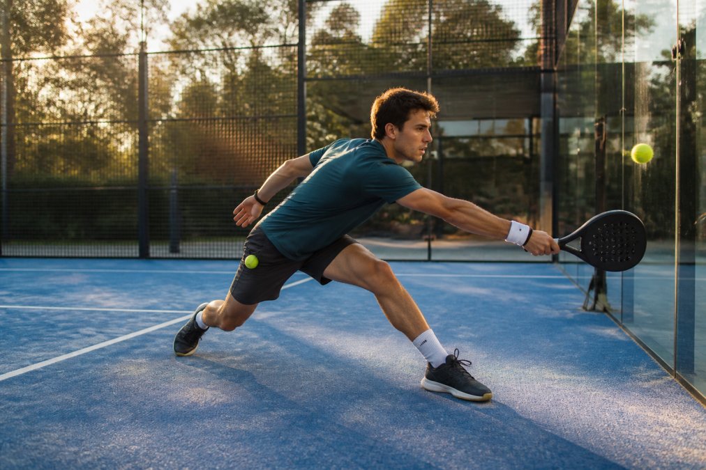 A padel player in breathable sportswear mid-lunge on an outdoor court, racket extended toward the glass wall, a spare ball visible in a side pocket, late afternoon sunlight