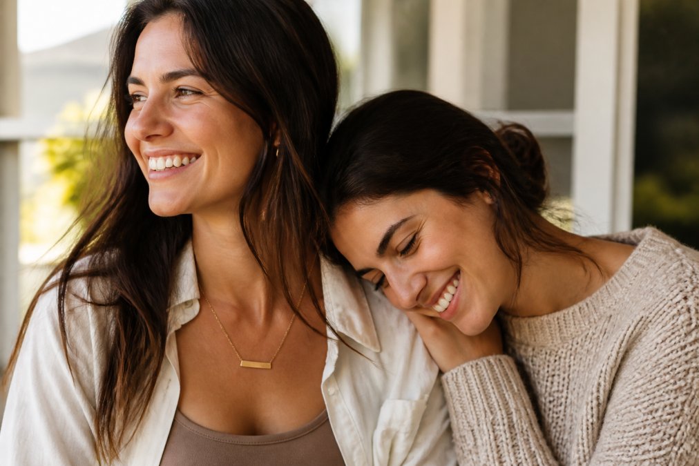 Close-up of two sisters smiling together, one wearing a delicate personalized name necklace, warm natural light, candid moment