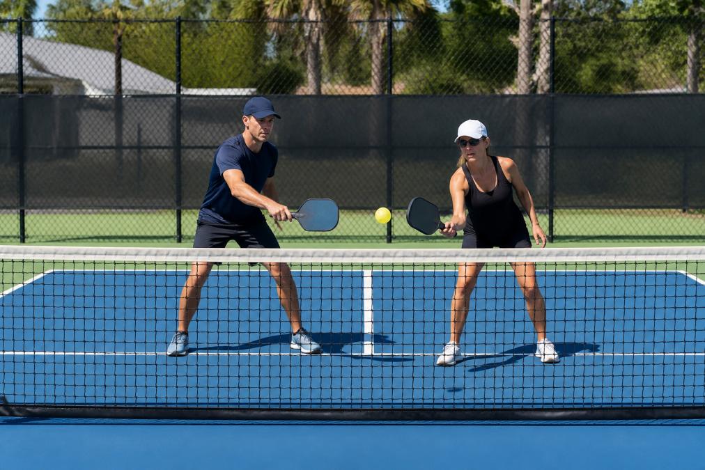 Pickleball players at the net during a dinking exchange, demonstrating control-focused play with proper court positioning and paddle angles in natural daylight