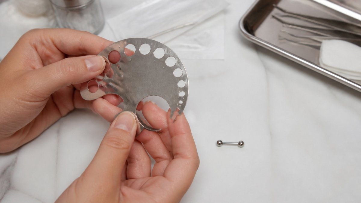 Hands using a sterile gauge to measure a small titanium barbell on a clean white surface