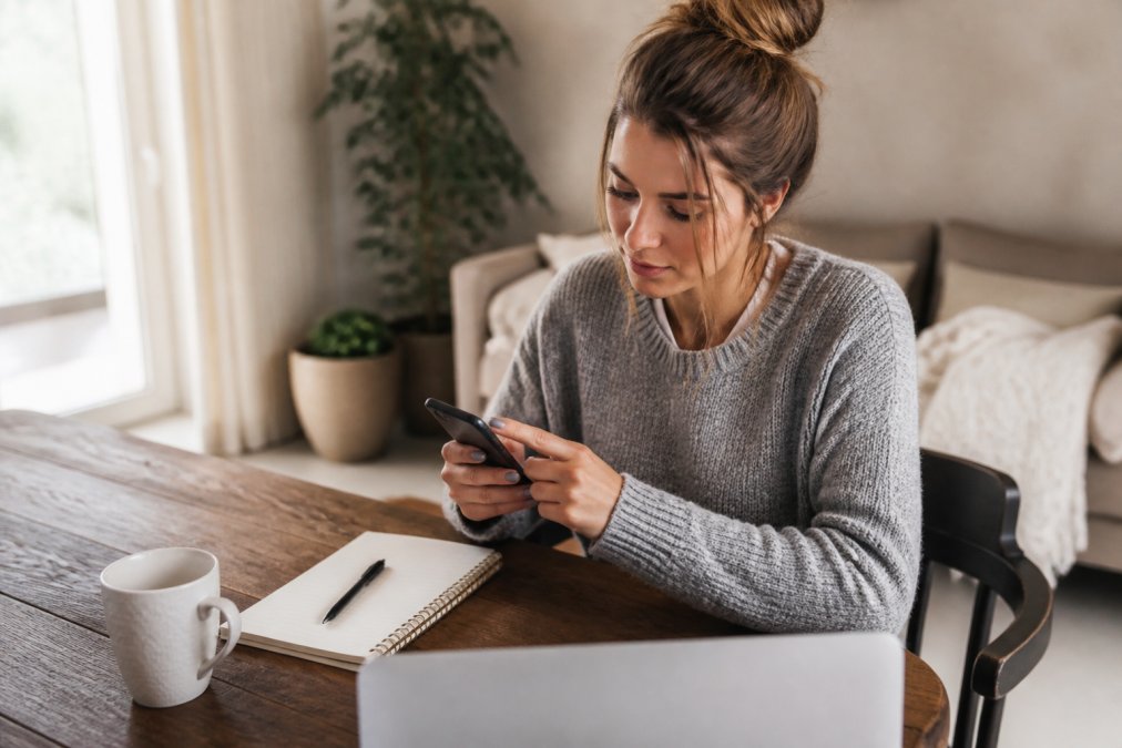 Woman at home office desk using smartphone to research Instagram content, with notebook and coffee cup visible, natural window lighting