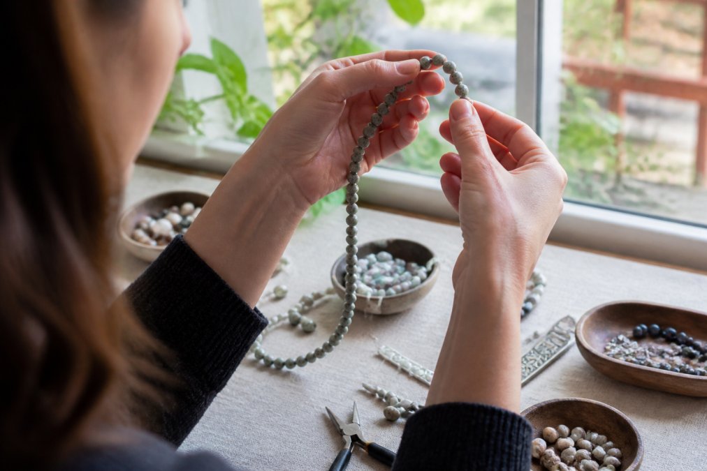 Close-up of a crafter's hands holding a strand of natural stone beads up to a bright window, examining a single bead for clarity and surface flaws