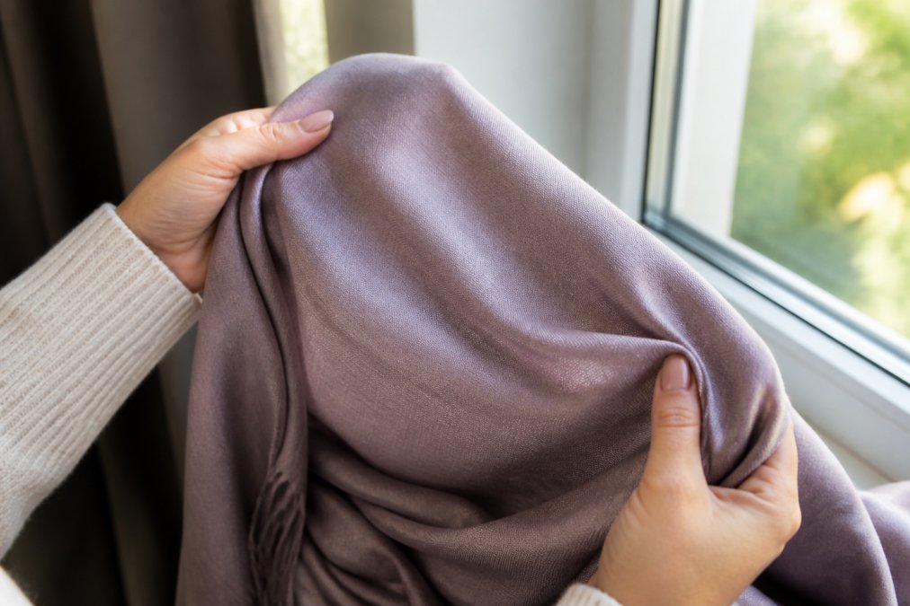 Close-up of hands holding a soft pashmina shawl up to natural window light, showing the fabric's drape and gentle sheen