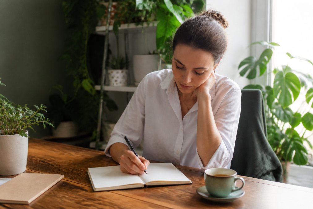 Woman writing goals in a notebook for reliable self-improvement in a bright, plant-filled home office workspace.