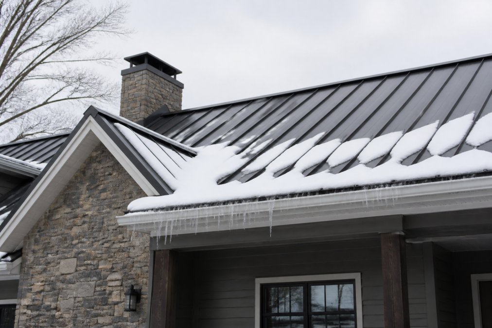 Residential home with standing seam metal roofing during winter conditions showing snow coverage clinging to the pitched roof surface, with ice formation visible at the eaves and gutters, set against a gray winter sky