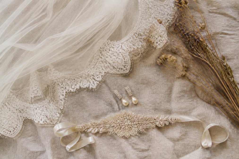 A flat lay of a vintage-inspired bridal ensemble including a cathedral veil, pearl drop earrings, and a delicate sash belt arranged on soft linen with dried flowers
