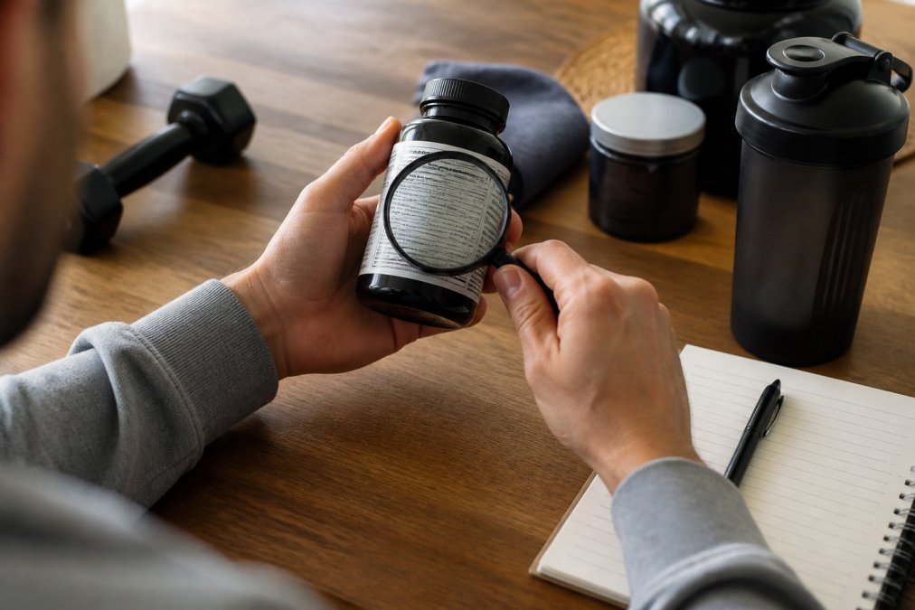 Close-up of hands holding a supplement bottle and examining the label with a magnifying glass next to a notepad on a clean desk