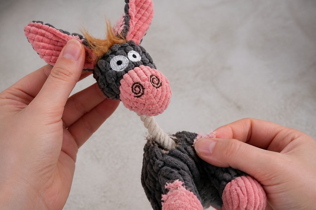Pet owner's hands carefully inspecting a worn dog toy for loose threads, frayed edges, and material deterioration against a neutral background