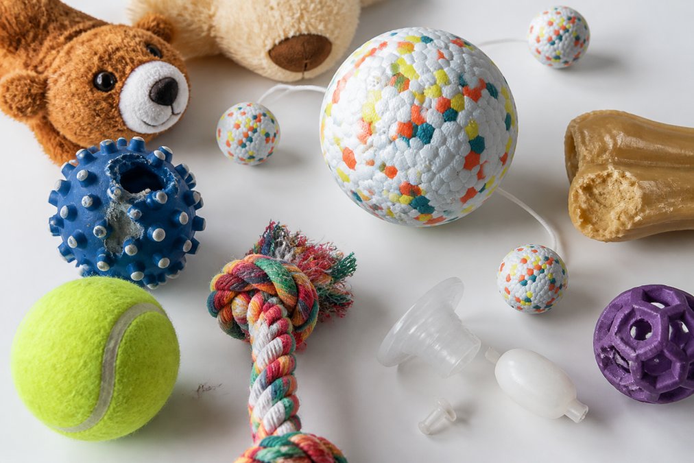 Close-up of various pet toys laid out on a white surface showing different materials, textures, loose threads, squeaker mechanisms, and small detachable parts under bright natural lighting