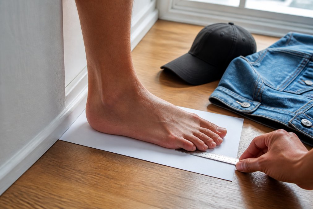 Close-up of a person's foot positioned on white paper against a wall with a ruler measuring from heel to longest toe, natural daylight from window