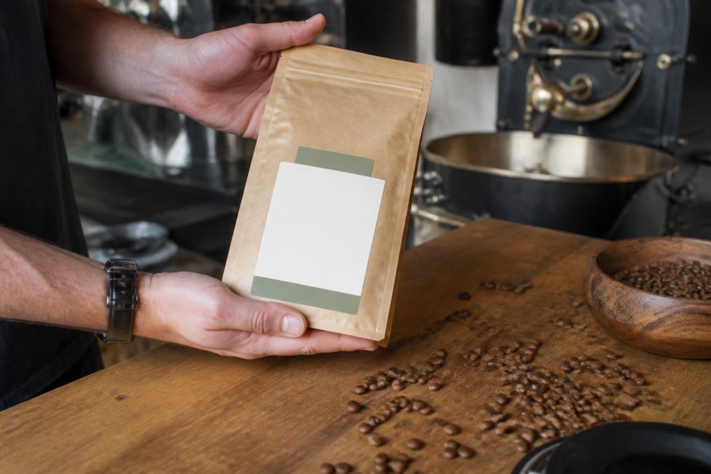 Close-up of hands holding a bag of specialty coffee beans, turning the label toward the camera to show origin and roast date details, with a few loose beans scattered on a wooden counter