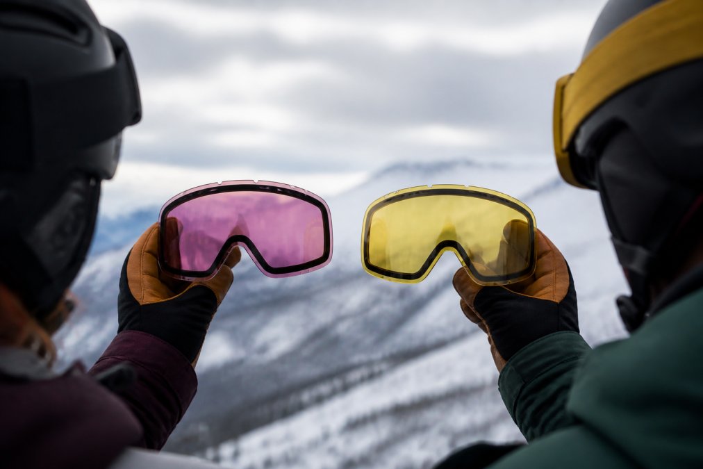 Close-up of a skier's gloved hands holding two interchangeable goggle lenses side by side, one rose-tinted and one yellow-tinted, with a snowy mountain slope blurred in the background under overcast skies