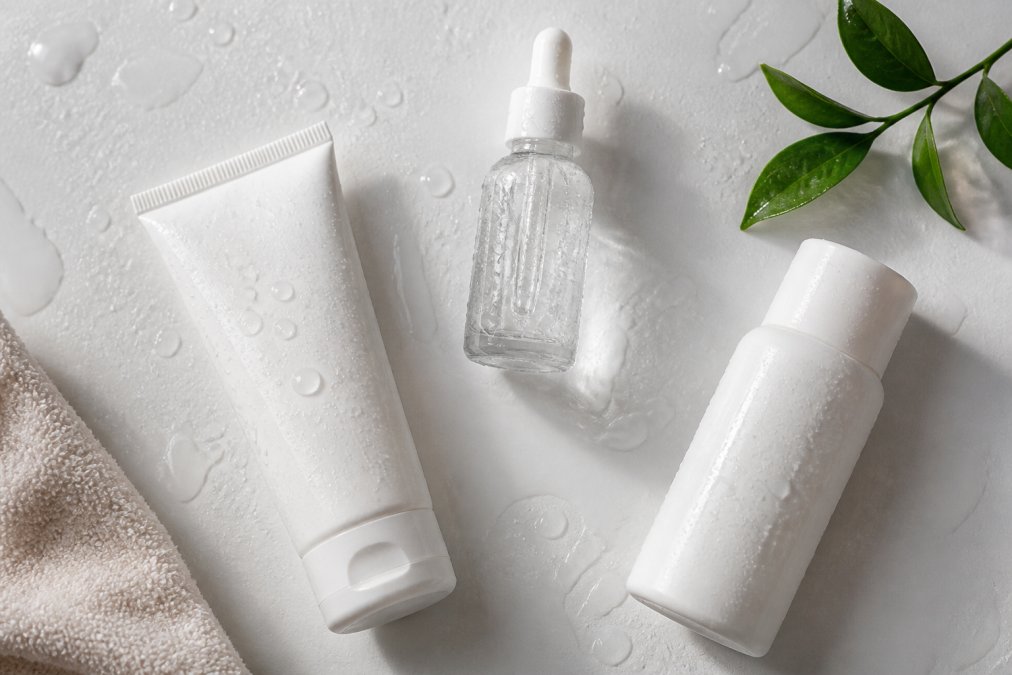 Flat lay of gentle skincare products including a mild cleanser, hydrating moisturizer with hyaluronic acid, and broad-spectrum sunscreen arranged on white surface with water droplets