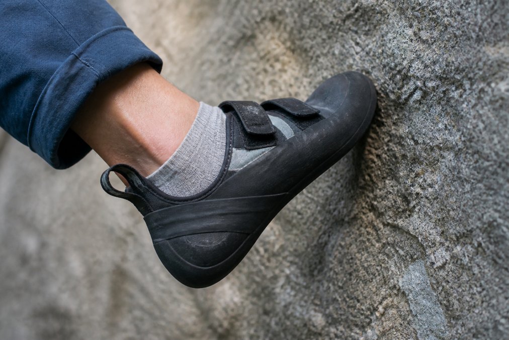 Close-up of a climber's foot positioned in a climbing shoe with a thin sock, showing precise toe box contact and heel tension against the rock wall