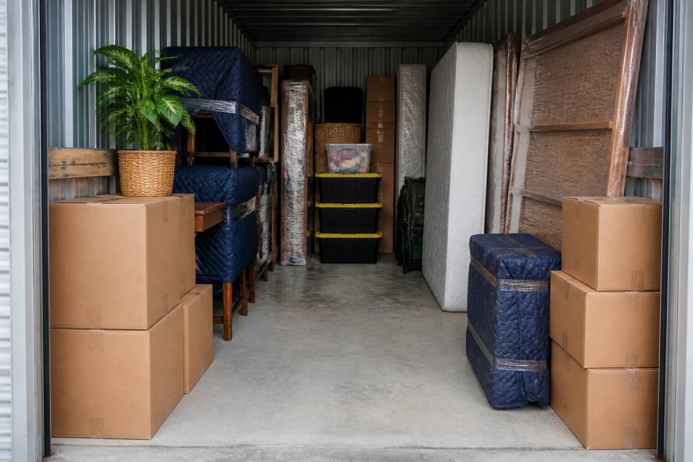 Interior view of a storage unit with neatly organised furniture stacked vertically, boxes arranged along walls, and clear aisle space for access