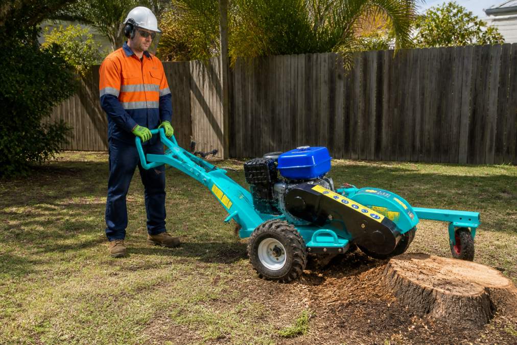 Operator wearing full safety gear including hard hat, face shield, eye protection, heavy-duty gloves, and steel-capped boots standing confidently behind a stump grinder in a residential backyard during daylight