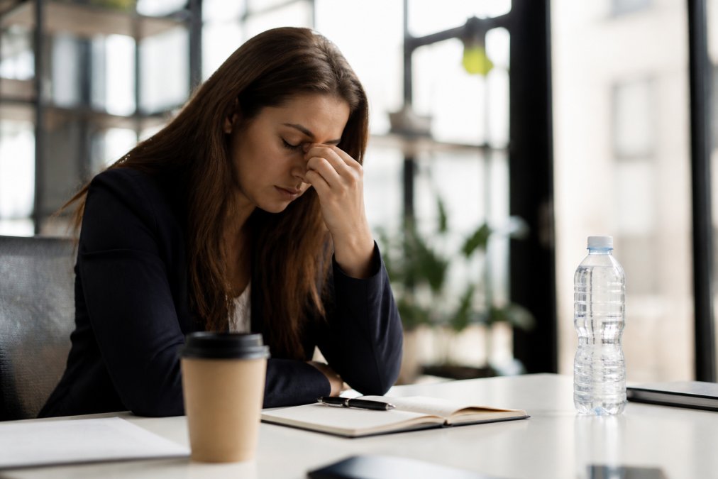Person looking visibly fatigued at desk during afternoon, with coffee cup and water bottle nearby, slumped posture showing signs of low energy