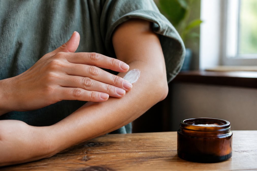 A close-up of a person's hands applying a small amount of white lotion to their forearm, with an amber glass product jar visible on a wooden table nearby, soft natural window lighting