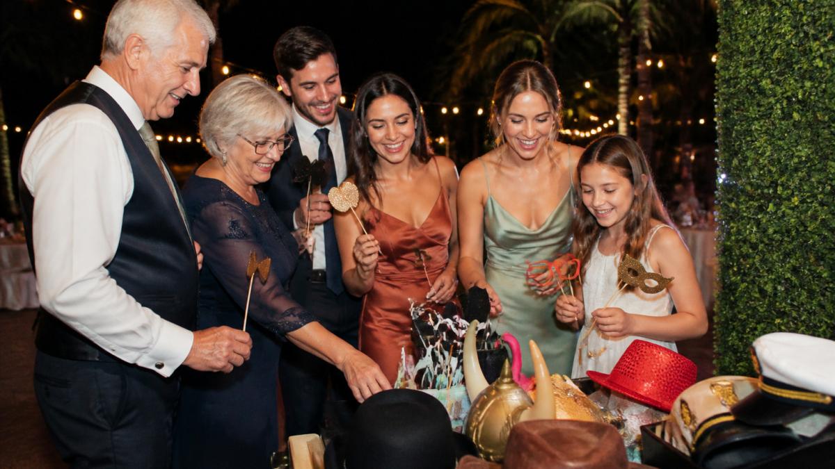 Wedding guests laughing while choosing fun photo booth props at an evening reception backdrop
