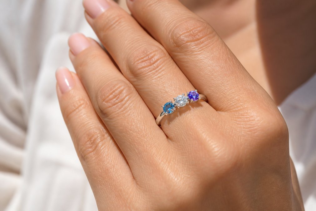 Close-up of an elegant mother's ring featuring three gemstones in white gold, displayed on a woman's hand with natural sunlight highlighting the sparkle and arrangement of the stones