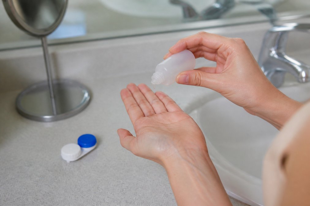 Close-up of a person's hands rinsing a contact lens with solution over a clean bathroom sink, with a lens case and solution bottle visible on the counter