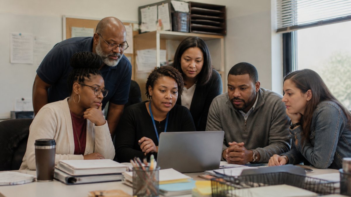 Diverse group of school staff and parent volunteers gathered around a laptop, reviewing a fundraising campaign dashboard together in a school office setting with natural afternoon lighting