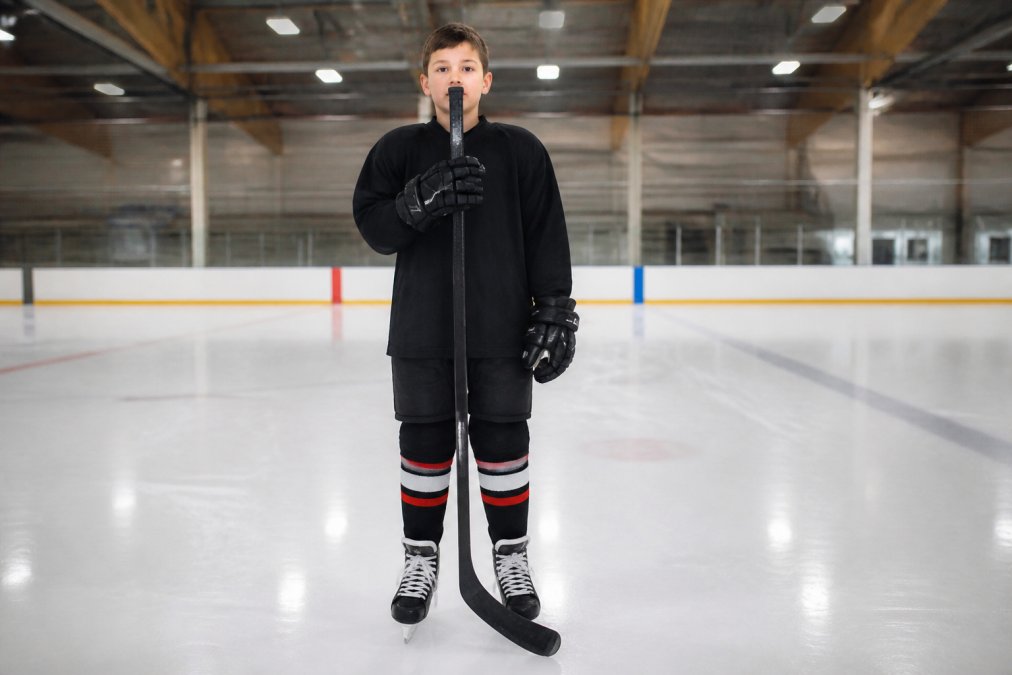 Young hockey player standing upright holding a hockey stick vertically from skate blade to chin, demonstrating proper measurement technique in indoor rink setting