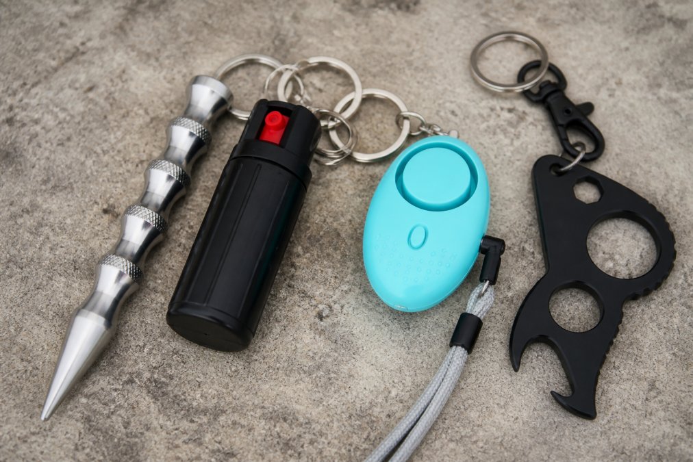 Close-up of various self-defense keychains displayed together on a neutral surface, including a metallic kubotan, a small pepper spray canister, a bright safety alarm, and a tactical keychain with multiple attachment points, showing size and design variety in natural daylight