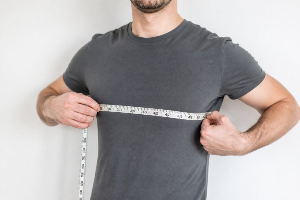 A person wrapping a flexible fabric tape measure around their chest at armhole level, wearing a thin fitted t-shirt, standing against a plain white wall in bright natural light
