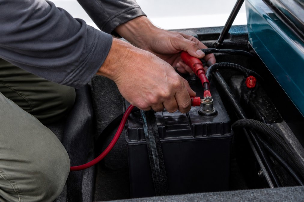 An angler's hands connecting a red battery cable to a terminal on a deep cycle marine battery inside a clean, organized boat battery compartment with natural daylight