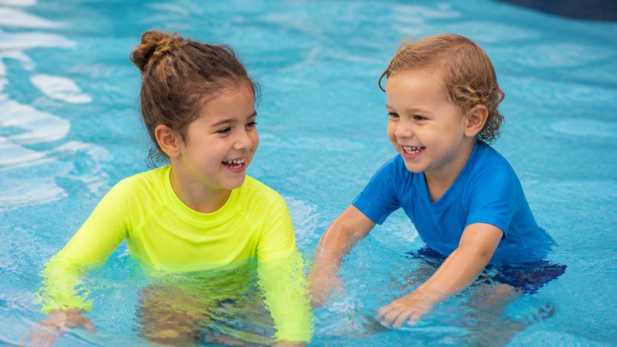 Two children of different ages playing together in a pool, one wearing a bright neon long-sleeve rash guard and the other in a short-sleeve option, both smiling and clearly visible against the water