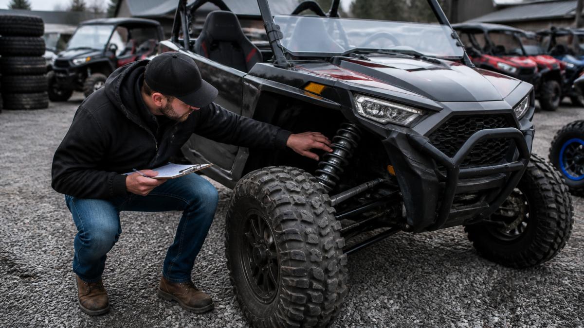 A person crouching next to a side-by-side UTV on a gravel lot, inspecting the front suspension and tire tread, with a clipboard in hand and a dealership lot in the background