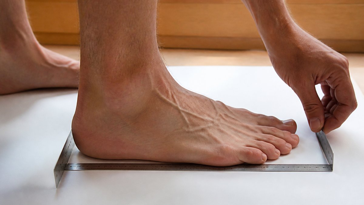 Close-up of a barefoot runner's foot being measured with a ruler on a flat white surface, showing proper heel-to-toe measurement technique with natural daylight