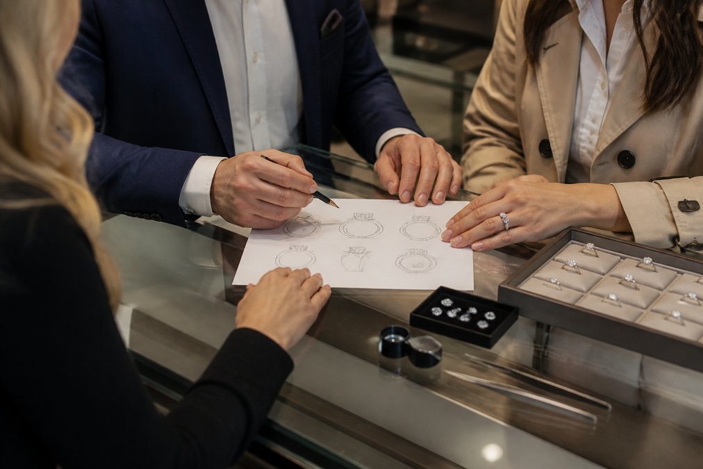A jeweler and a couple reviewing hand-drawn ring sketches and loose gemstones at a well-lit design counter, with a loupe and tweezers visible on the work surface
