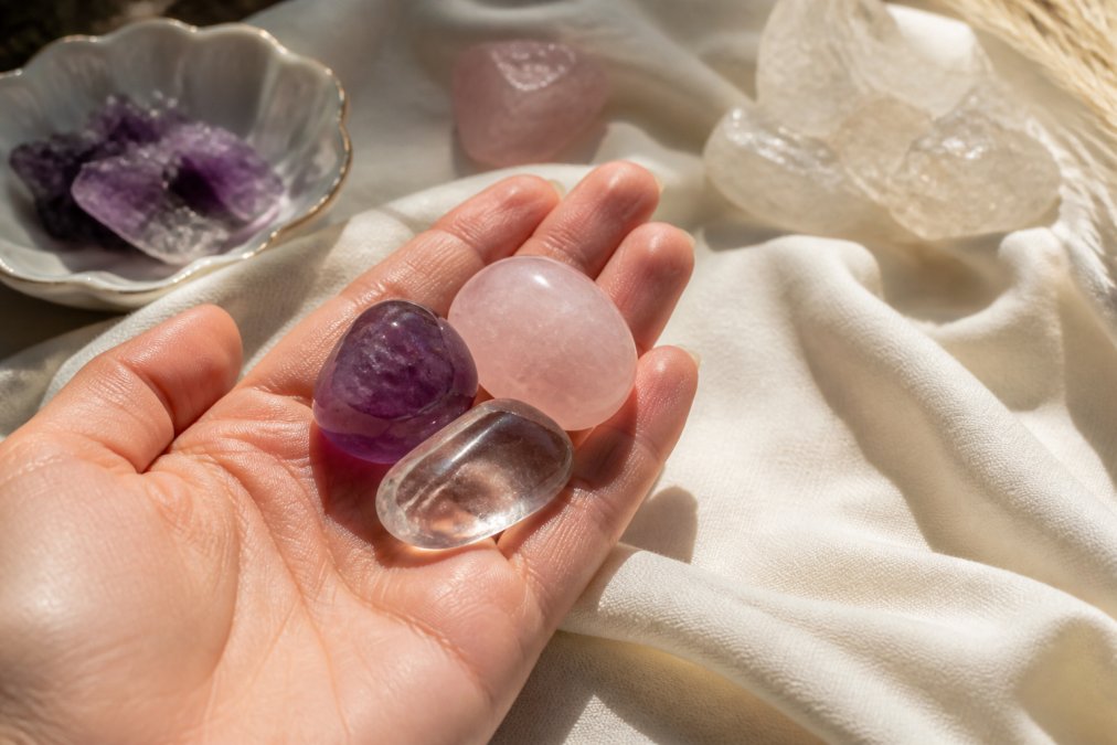 Close-up of a hand holding several polished beginner crystals including amethyst with purple coloring, rose quartz in soft pink, and clear quartz in transparent clarity, arranged on a cream-colored fabric with warm natural window lighting
