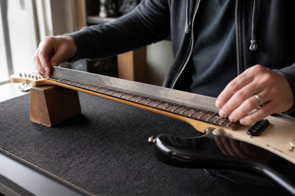Close-up of hands inspecting an electric guitar neck against natural light, checking for straightness and fret condition with a straightedge tool